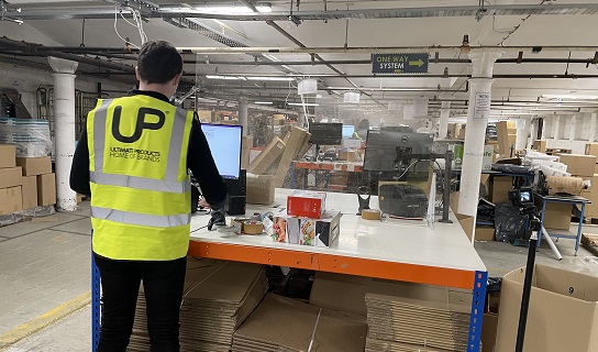 An image showing a person standing in front of a packing bench in a warehouse. They are looking at a computer screen and there are products on top of the bench.