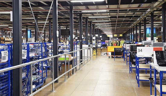 An image showing a a warehouse with wooden pack benches along the right hand side and picking trolleys containing products on the left hand side